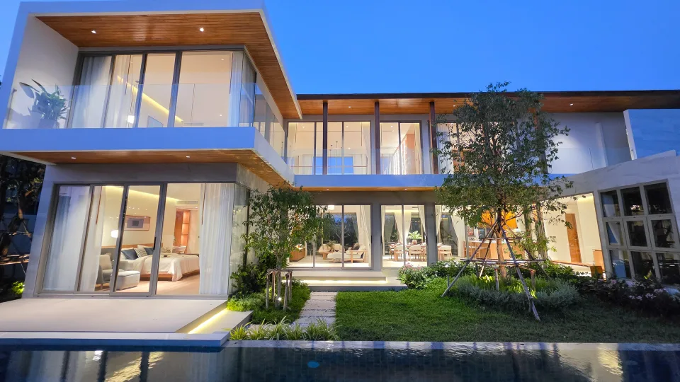 Two-storey villa lit at dusk, seen across the pool with the living area and bedroom open to the garden