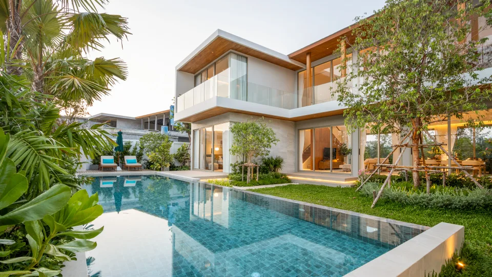 Villa exterior in daylight, seen across the pool with sun loungers on the far side