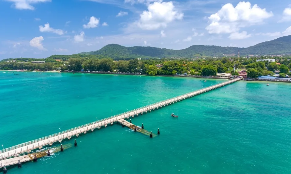Rawai Pier and beach in Phuket, Thailand, showcasing a long concrete pier extending into the crystal-clear turquoise ocean. The surrounding landscape features a lush green coastline with rolling hills in the background under a bright blue sky dotted with fluffy white clouds, highlighting a serene and picturesque tropical setting.